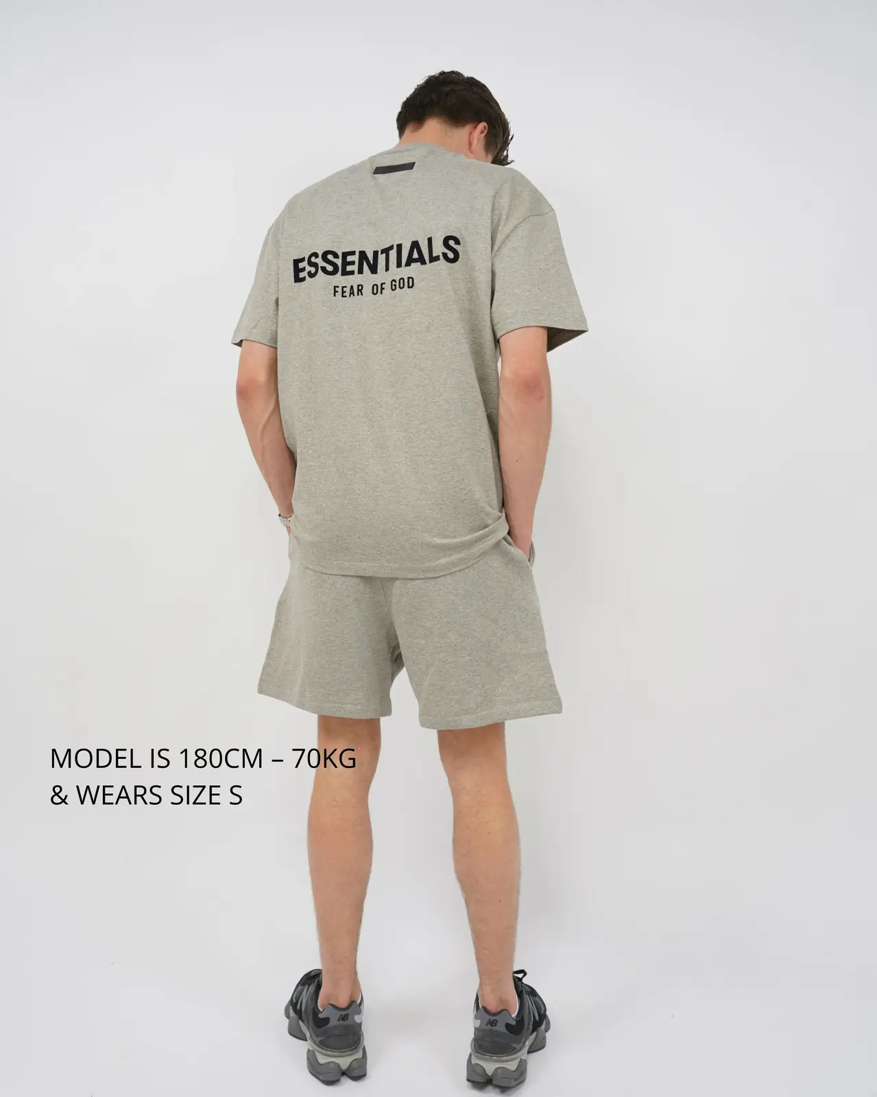 Rear view of a man wearing the Fear of God Essentials Dark Oatmeal Summer set with a loose-fitting short-sleeve top and matching shorts, paired with black sneakers, against a plain white background.