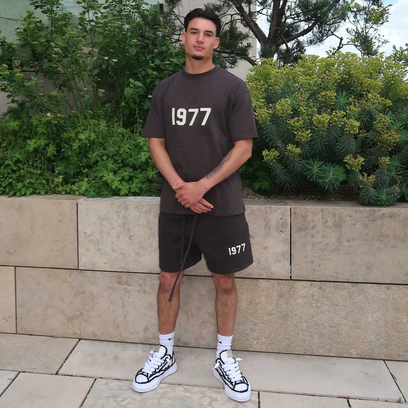 Man wearing dark gray Fear of God Essentials 1977 Iron Summer Set with 1977 printed on both shirt and shorts, paired with white sneakers against a greenery backdrop.