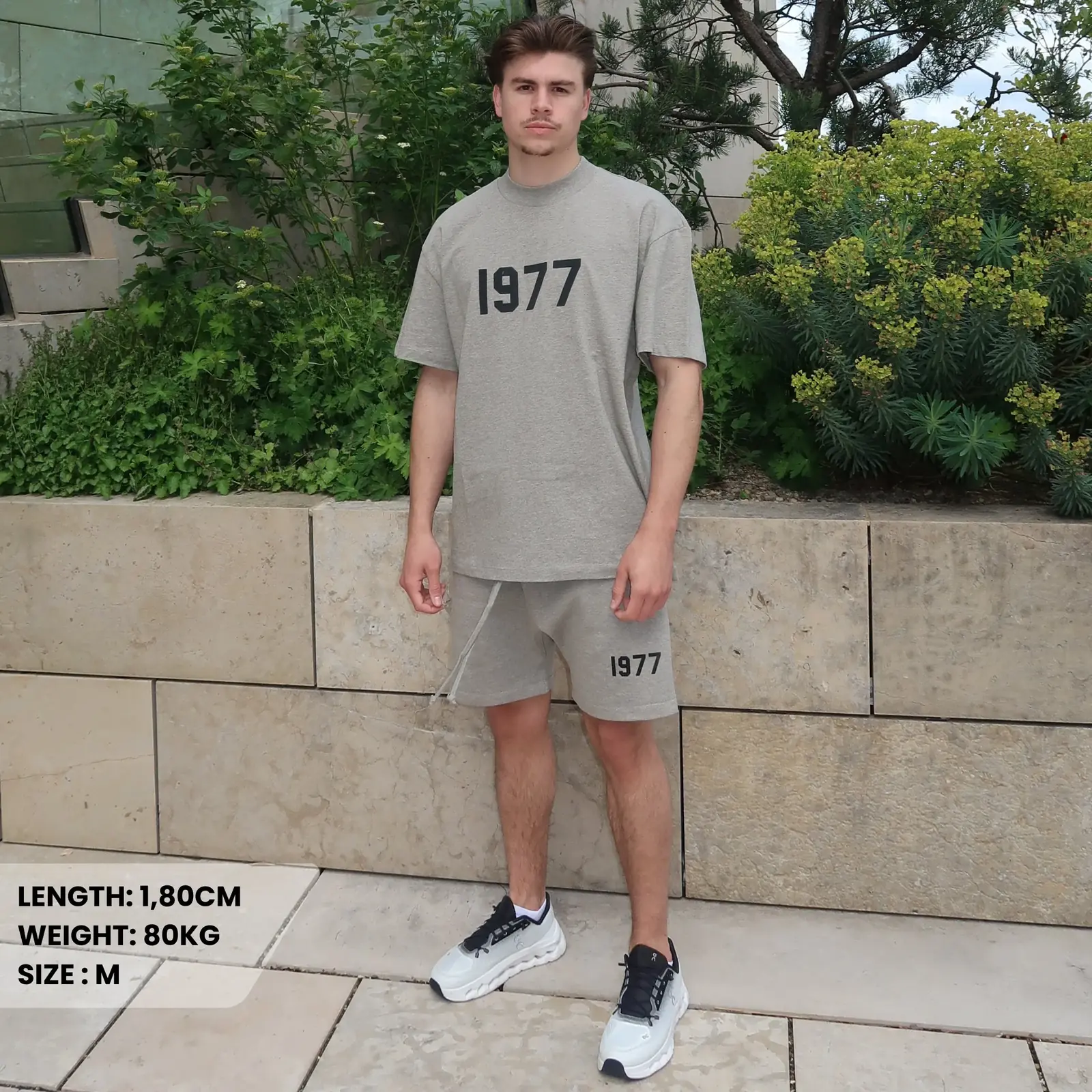 A young man wearing the Fear of God Essentials 1977 Dark Oatmeal Summer Set consisting of a grey oversized t-shirt and matching shorts with "1977" printed on both, paired with white and black sneakers, standing outdoors by a stone wall and greenery, showcasing exclusive European style.