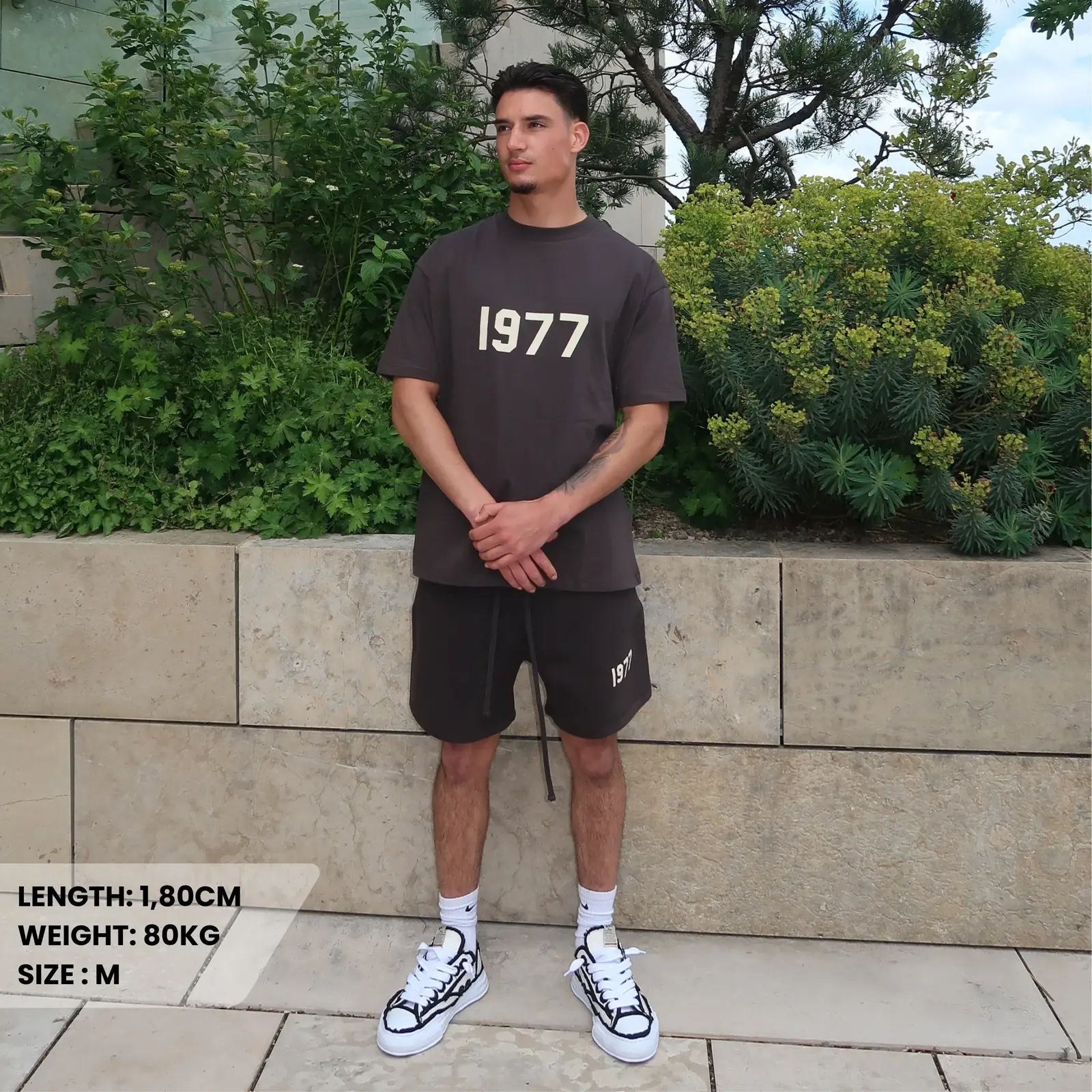 Man wearing Fear of God Essentials 1977 Iron Summer Set in dark gray, featuring a 1977 print on both shirt and shorts, paired with white sneakers, standing outdoors near greenery, The real deal.