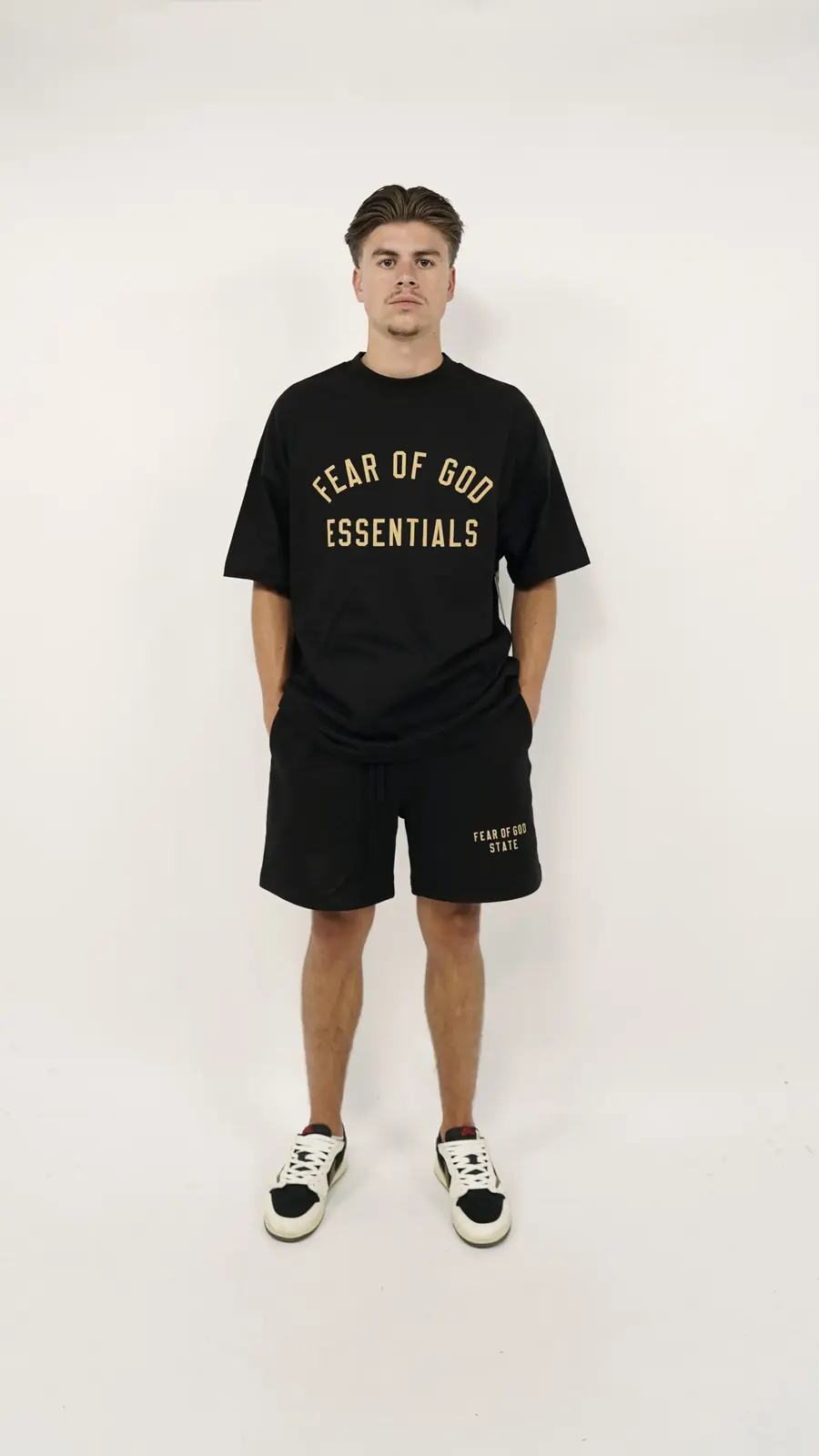 A man wearing the Fear of God Essentials FW24 Black Yellow Summer Set consisting of a black oversized t-shirt with yellow lettering and matching black shorts, paired with black and white sneakers, standing against a plain white background.