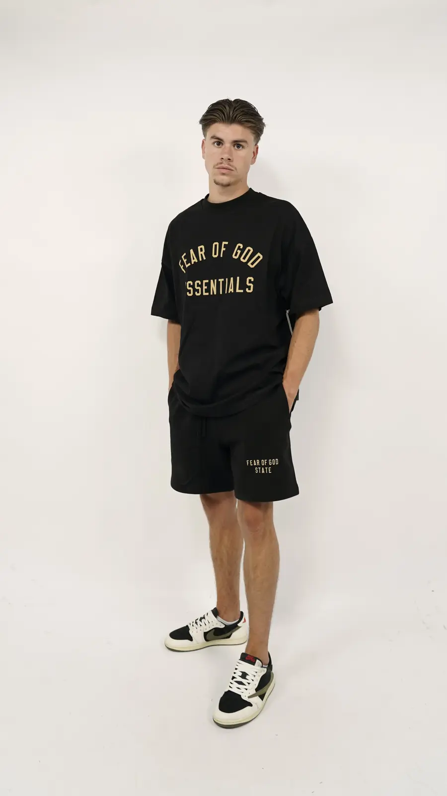 Man wearing Fear of God Essentials FW24 Black Yellow Summer Set with black oversized shirt and shorts paired with black and white sneakers, minimalist studio background.