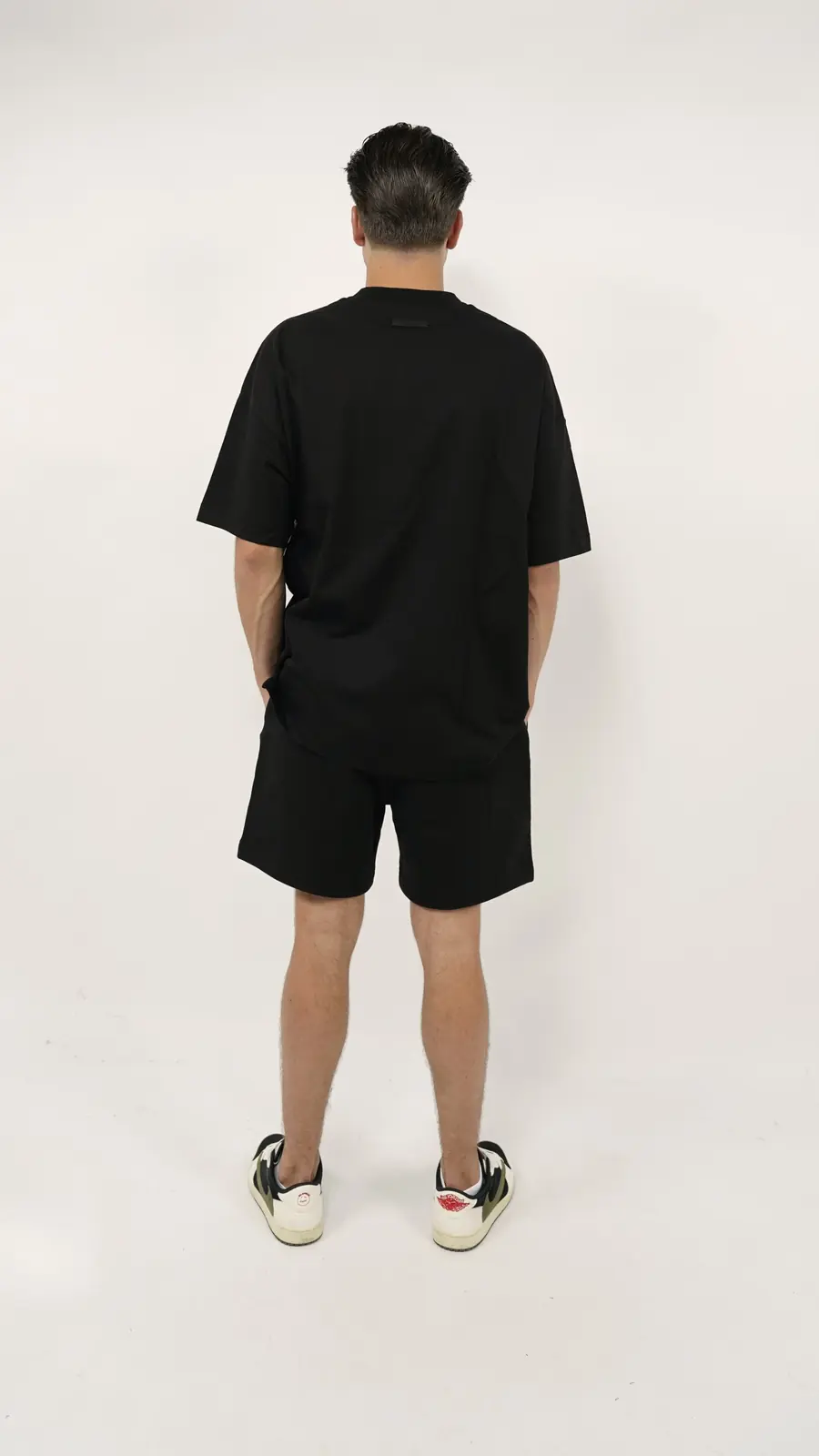 Man seen from behind wearing the Fear of God Essentials FW24 Black Yellow Summer Set with black oversized t-shirt, matching shorts, and black and white sneakers, showcasing a clean and minimalist summer look.