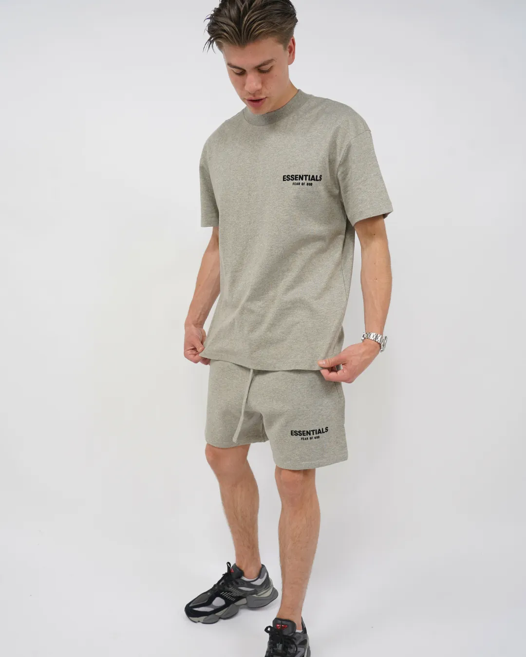 A young man wearing a Fear of God Essentials Dark Oatmeal Summer set with a comfortable short-sleeve top and matching shorts paired with casual black sneakers, showcasing a relaxed summer outfit.