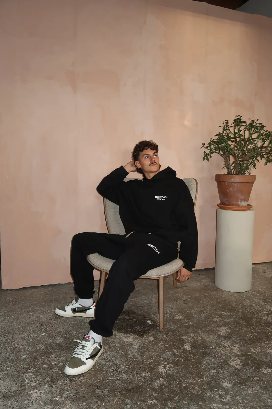 Man sitting on a chair wearing Fear of God Essentials FW24 Black White Lange Broek with black hoodie and white sneakers, in a minimalist room with a potted plant and textured floor, showcasing exclusive sneakers in whole Europe. The real deal.
