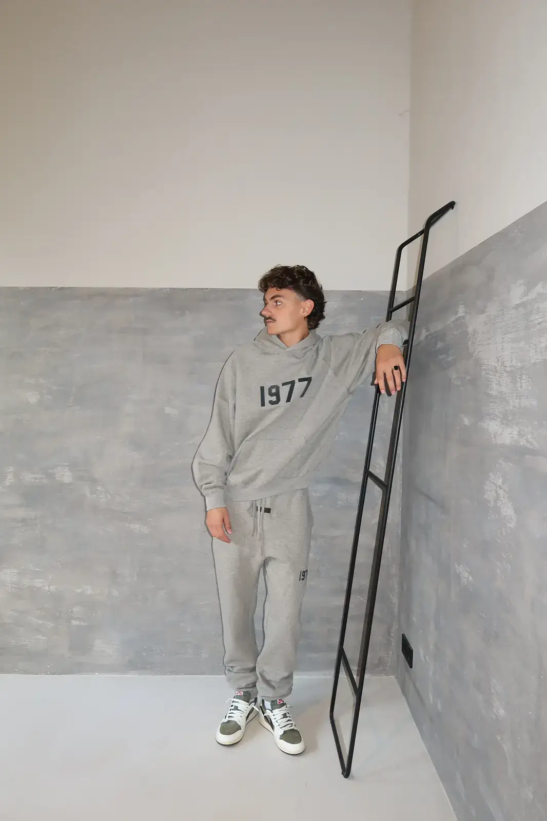 Man wearing Fear of God Essentials 1977 Dark Oatmeal Lange Broek sweatpants and matching hoodie, posing casually against a modern concrete wall while leaning on a black ladder, showcasing exclusive clothing style.