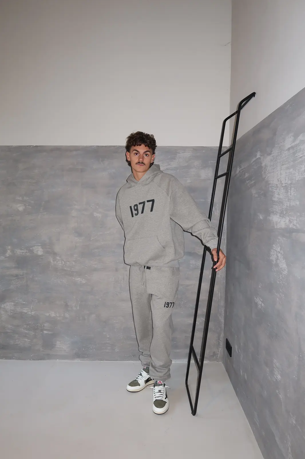 Man wearing a gray matching hoodie and pants set with "1977" printed on both pieces, paired with sneakers, standing indoors holding a black metal ladder, showcasing Fear of God Essentials 1977 Dark Oatmeal Lange Broek.