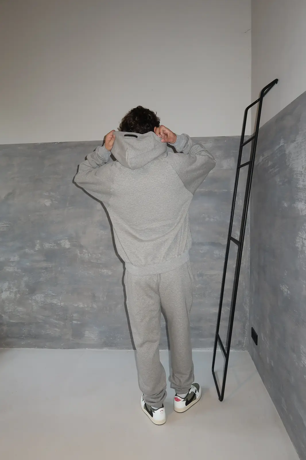 Person wearing the Fear of God Essentials 1977 Dark Oatmeal Full set, shown from the back in a minimalist setting with gray walls and casual sneakers, embodying The real deal.