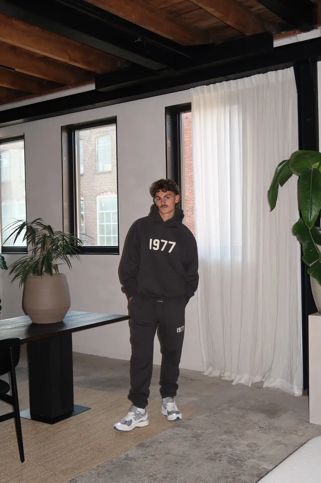 A man indoors wearing a black Fear of God Essentials 1977 Iron Black Lange Broek tracksuit with white "1977" lettering on hoodie and pants, paired with gray sneakers, standing near a window with a plant and natural light, representing exclusive sneakers in Europe, the real deal.