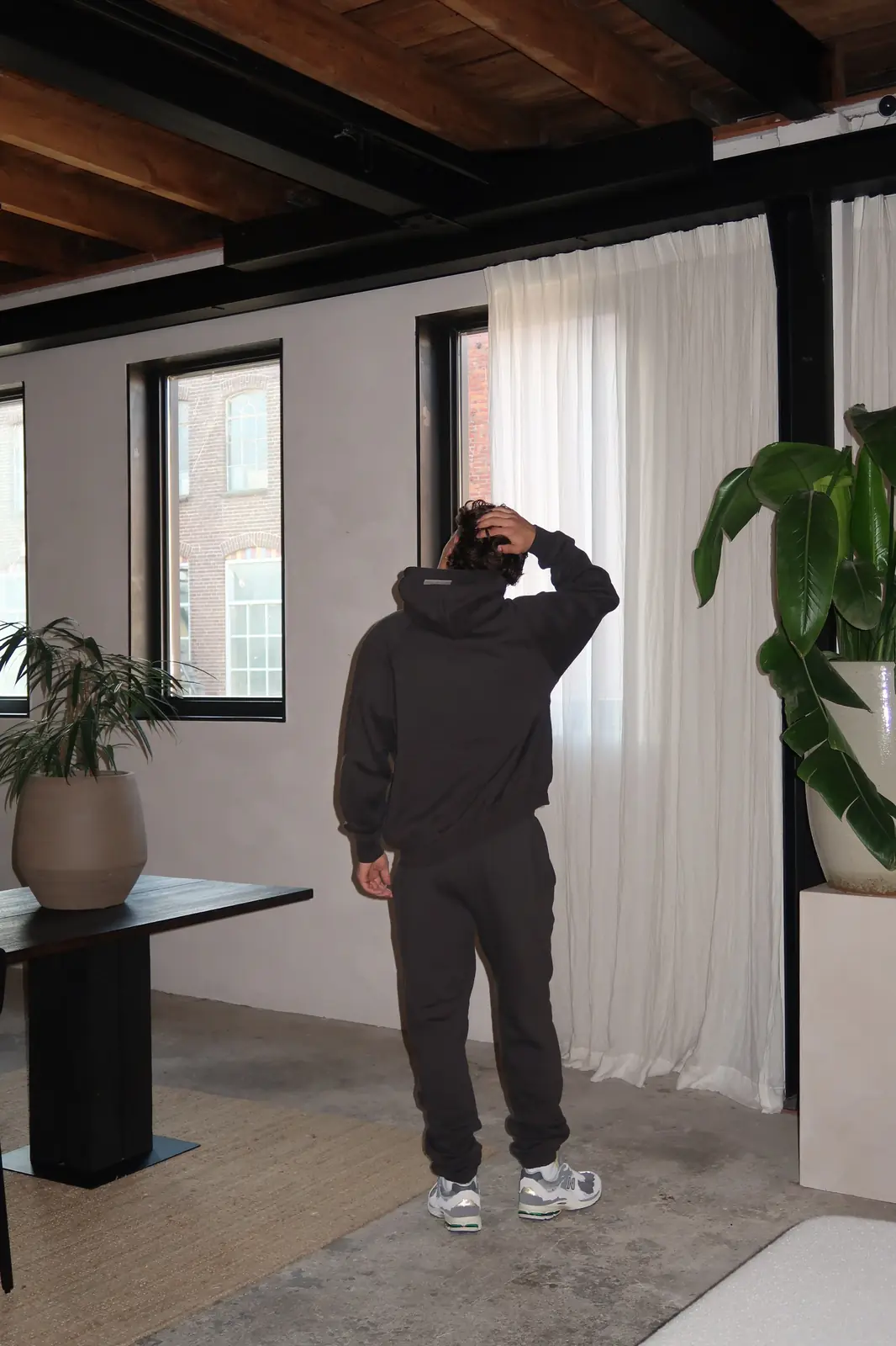 Man wearing Fear of God Essentials 1977 Iron Black Lange Broek standing indoors with hand on head, modern casual outfit paired with sneakers, urban minimalist setting.