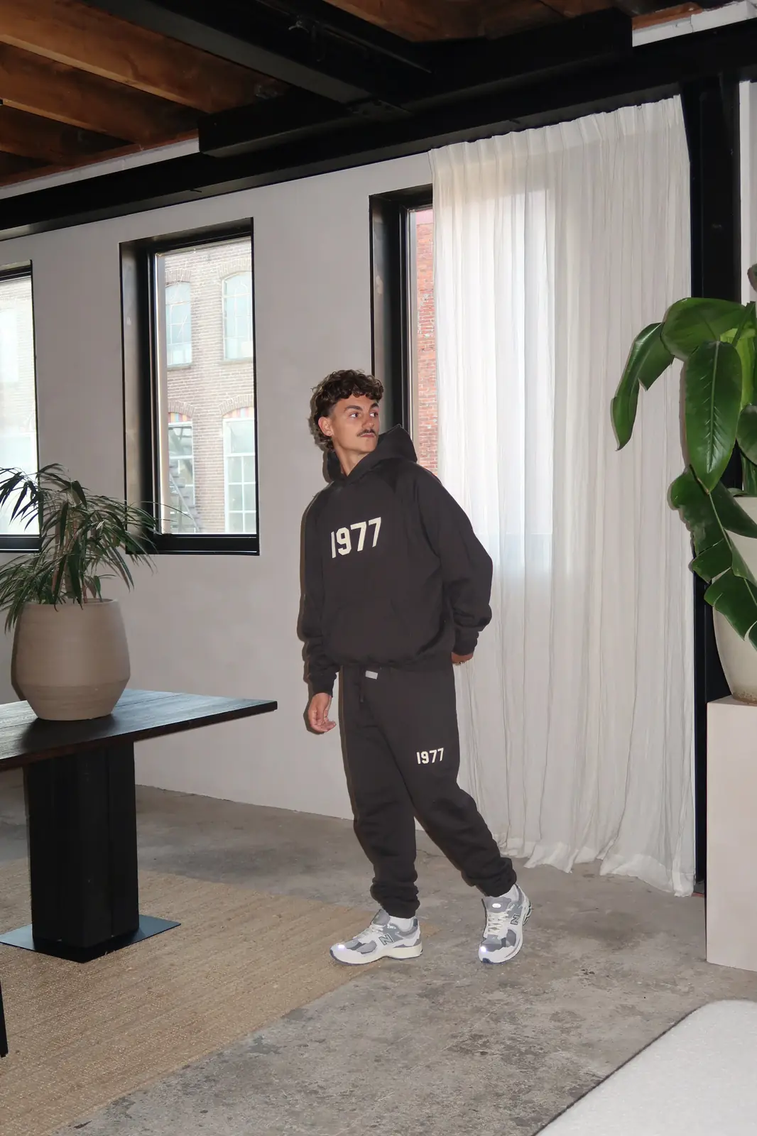 A man indoors wearing a Fear of God Essentials 1977 Iron Black Lange Broek with a matching hoodie, styled casually with white and gray sneakers, embodying The real deal.