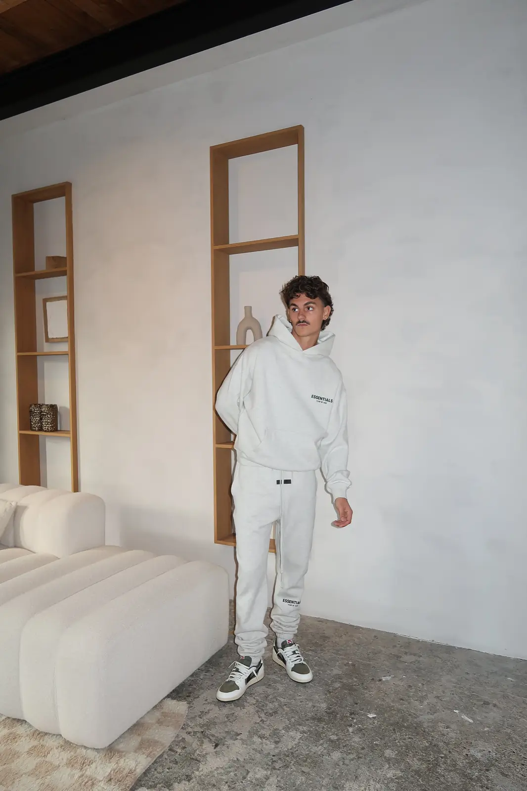 Man wearing a Fear of God Essentials Light Oatmeal Hoodie paired with matching sweatpants and sneakers, standing in a minimalist room with neutral tones, showcasing casual streetwear style.