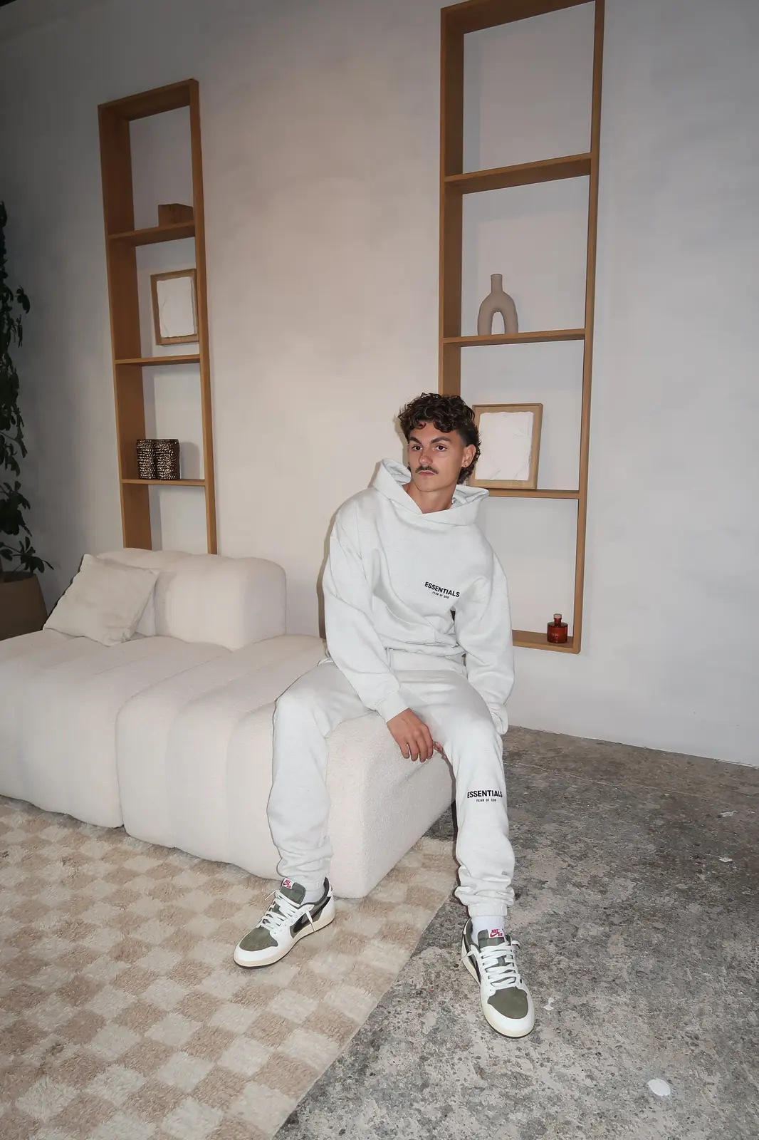 Young man wearing Fear of God Essentials Light Oatmeal Lange Broek seated on a modern cream sofa in a minimalist living room with neutral tones and wooden shelving.