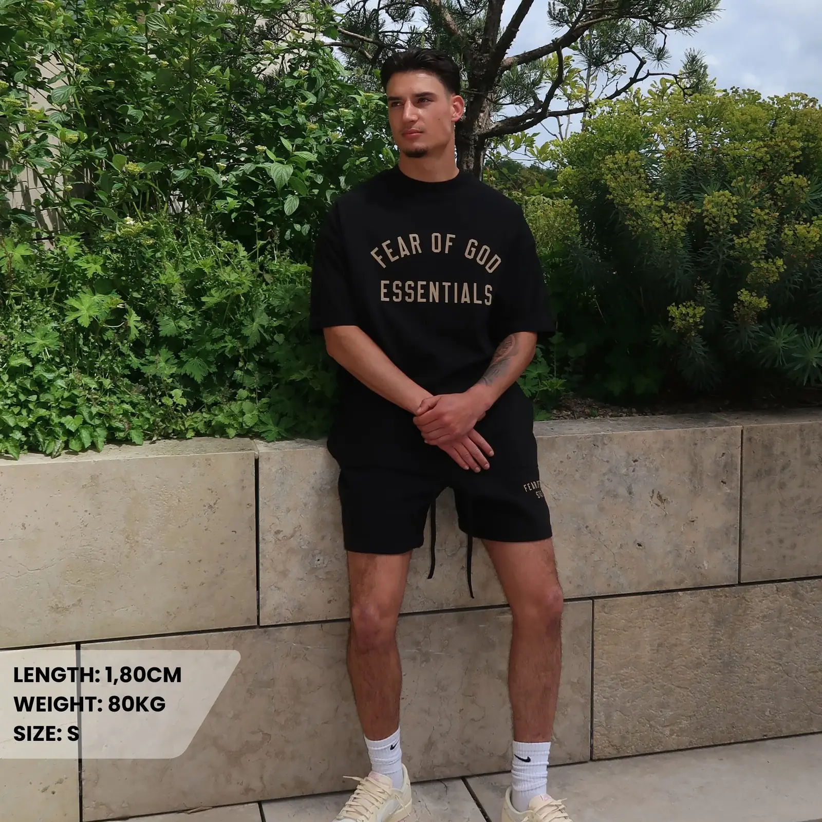 Man wearing Fear of God Essentials FW24 Black Yellow Summer Set with black t-shirt and shorts posing outdoors in casual sneakers, surrounded by greenery.