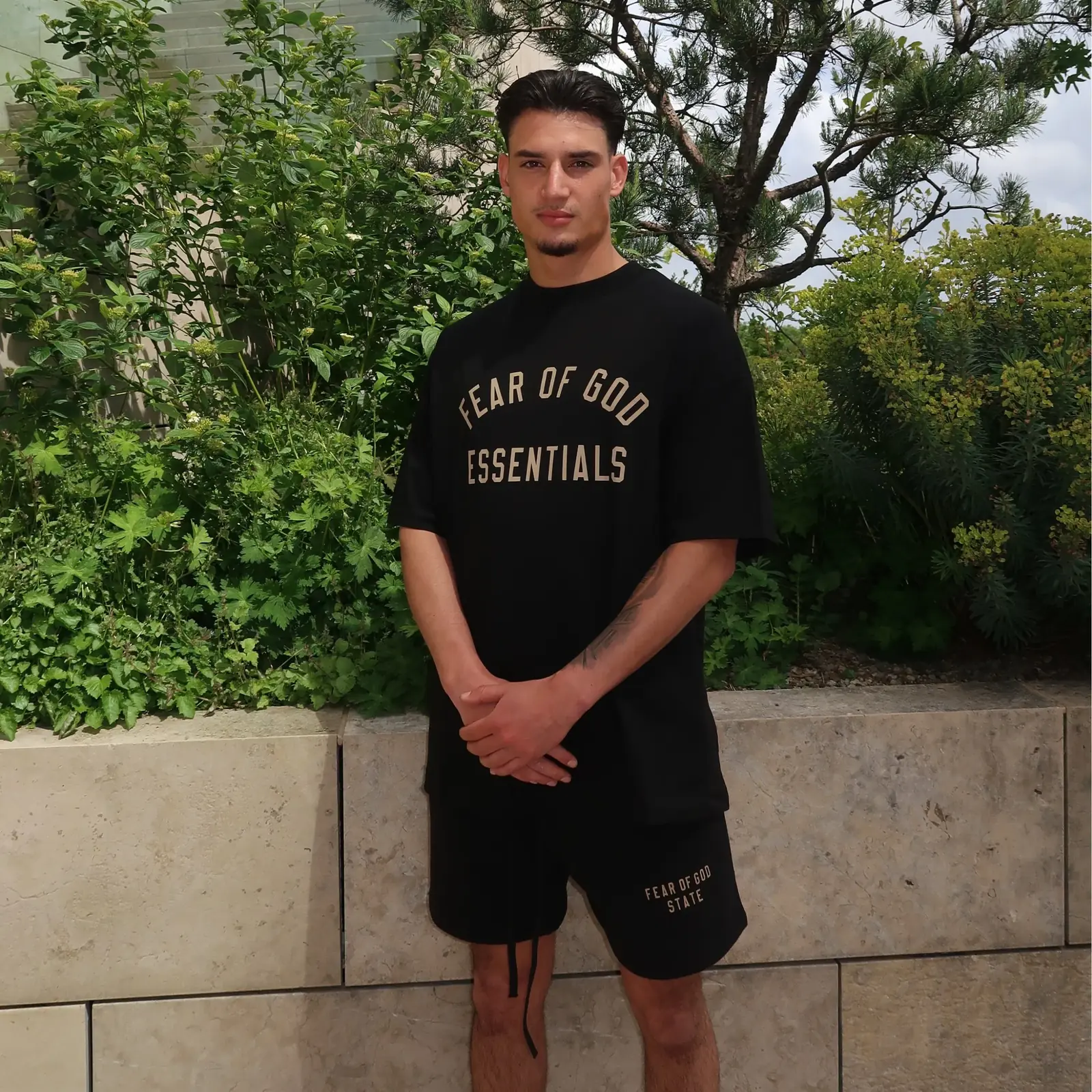A man outdoors wearing the Fear of God Essentials FW24 Black Yellow Summer Set, featuring a black oversized t-shirt and matching shorts with beige lettering, showcasing casual summer clothing.