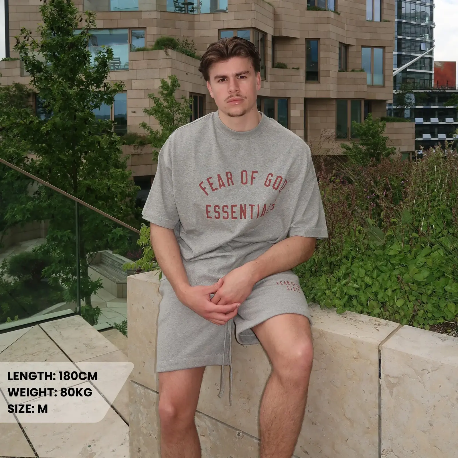 Man sitting outdoors wearing a grey Fear of God Essentials FW24 Dark Heather Summer Set with matching shorts and T-shirt embroidered with red lettering, promoting exclusive sneakers in Europe.