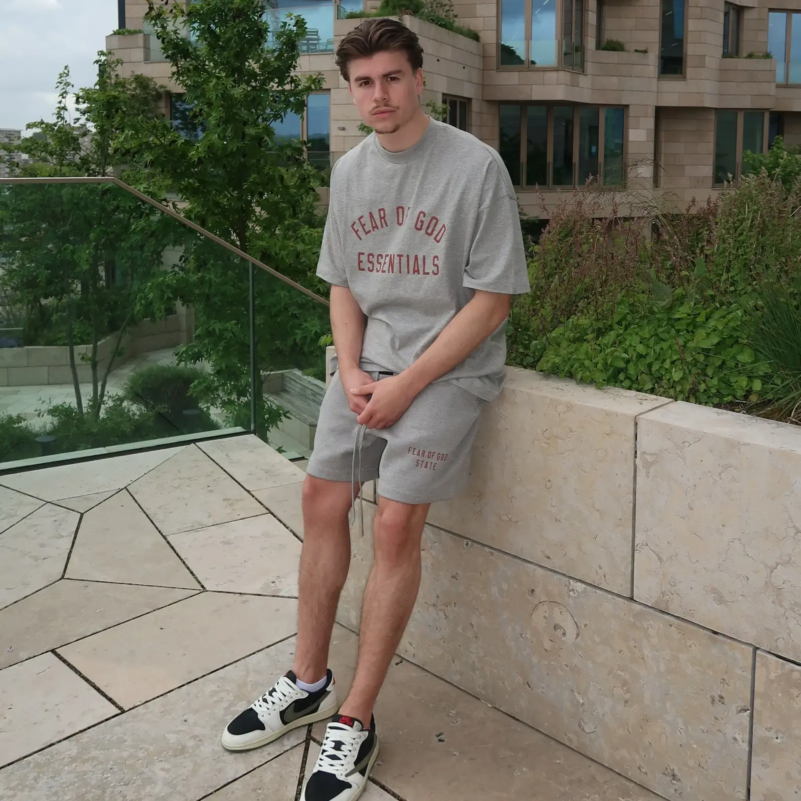 A young man wearing the Fear of God Essentials FW24 Dark Heather Summer Set in gray, paired with stylish sneakers, sitting outdoors on a stone bench with greenery in the background.
